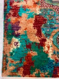 Azerbaijan Tappeti Milano Primo piano di un tappeto colorato con motivi astratti in rosso, blu, verde acqua e beige, con trama intrecciata visibile e frange sul bordo.