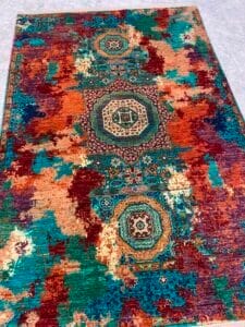 Azerbaijan Tappeti Milano Su un pavimento piastrellato è steso un tappeto colorato con motivi geometrici e una fila centrale di medaglioni nei toni del verde acqua, dell'arancione e del marrone.