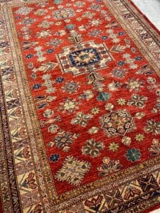 Azerbaijan Tappeti Milano Un tappeto orientale rosso con intricati motivi geometrici e floreali nei toni del blu, del beige e del verde, bordato da motivi dettagliati. Un logo multicolore a forma di farfalla appare nell'angolo.