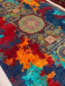 Azerbaijan Tappeti Milano Un tappeto colorato con motivi geometrici e floreali nei toni del blu, rosso, arancione e verde acqua, caratterizzato da medaglioni centrali e transizioni di colore pixelate.