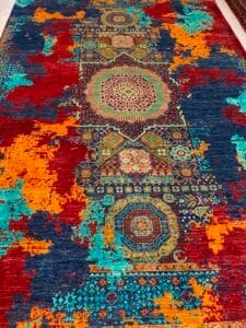 Azerbaijan Tappeti Milano Un tappeto colorato caratterizzato da intricati motivi geometrici con sezioni in rosso, arancione, blu e turchese e un logo a forma di foglia multicolore nell'angolo in alto a destra.