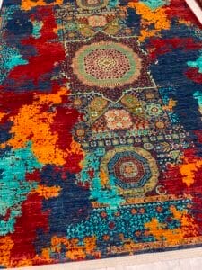 Azerbaijan Tappeti Milano Un tappeto colorato con un motivo geometrico centrale, caratterizzato da sezioni rosse, blu, arancioni e verde acqua e da un disegno astratto di schizzi di vernice.