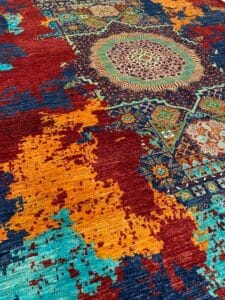 Azerbaijan Tappeti Milano Primo piano di un tappeto colorato con motivi geometrici e zone rosse, arancioni, blu, verdi e marrone chiaro.