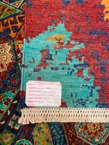 Azerbaijan Tappeti Milano Primo piano di un angolo di un tappeto colorato intrecciato con un'etichetta bianca che ne riporta dettagli quali origine, qualità, dimensioni e composizione, che copre parzialmente un altro tappeto decorato.