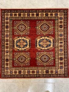 Azerbaijan Tappeti Milano Un tappeto quadrato rosso e beige con disegni geometrici è adagiato su un pavimento piastrellato di colore chiaro.