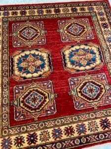 Azerbaijan Tappeti Milano Un tappeto orientale rosso con motivi geometrici e un bordo decorativo nei colori blu, oro e crema.