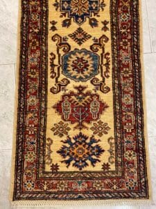Azerbaijan Tappeti Milano Un tappeto tradizionale con motivi geometrici nei toni del rosso, blu e oro su un pavimento piastrellato.