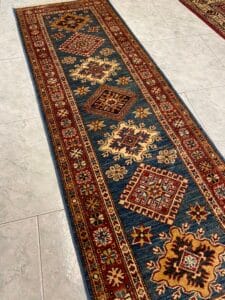 Azerbaijan Tappeti Milano Tappeto tradizionale con motivi geometrici in rosso, giallo e blu su pavimento piastrellato.