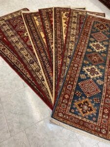 Azerbaijan Tappeti Milano Cinque tappeti con motivi geometrici tradizionali e diverse combinazioni di colori sono disposti uno accanto all'altro su un pavimento piastrellato.