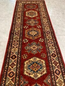 Azerbaijan Tappeti Milano Un lungo tappeto orientale rettangolare con motivi geometrici nei toni del rosso, beige, blu e verde su un pavimento piastrellato chiaro.