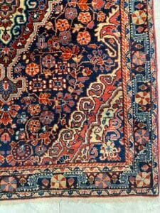 Azerbaijan Tappeti Milano Intricato e colorato tappeto persiano con motivi floreali e geometrici su pavimento piastrellato; logo a forma di farfalla in un angolo.