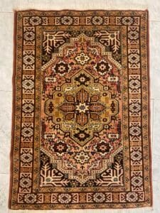 Azerbaijan Tappeti Milano Un tappeto rettangolare con motivi geometrici nei toni del marrone, del beige, del nero e del rosso, adagiato su un pavimento piastrellato.