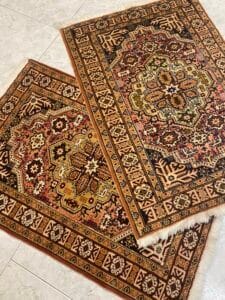 Azerbaijan Tappeti Milano Due tappeti orientali con motivi intricati e motivi geometrici, posizionati su un pavimento piastrellato e leggermente sovrapposti.