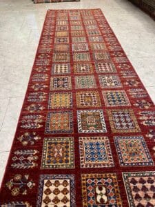 Azerbaijan Tappeti Milano Un lungo tappeto rosso con motivi geometrici quadrati in vari colori su un pavimento piastrellato.