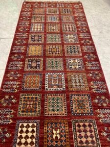 Azerbaijan Tappeti Milano Un tappeto rosso con colorati disegni geometrici quadrati e un logo multicolore a forma di foglia nell'angolo.