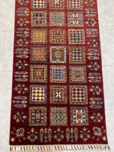 Azerbaijan Tappeti Milano Un tappeto rettangolare con motivi geometrici quadrati colorati disposti in file e sfondo rosso.