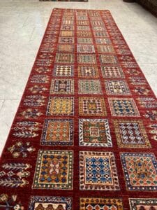 Azerbaijan Tappeti Milano Un lungo tappeto rosso decorato con quadrati geometrici colorati è steso su un pavimento piastrellato chiaro.