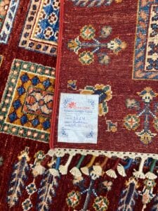 Azerbaijan Tappeti Milano Un tappeto con un motivo rosso, un angolo ripiegato e un'etichetta bianca con dettagli scritti a mano e nappe.