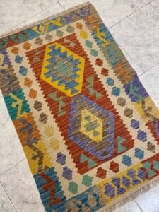 Azerbaijan Tappeti Milano Un tappeto intrecciato con motivi geometrici colorati a forma di rombi su un pavimento piastrellato chiaro.