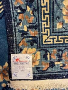 Azerbaijan Tappeti Milano Primo piano di un tappeto con motivo blu, un'etichetta di servizio bianca e un logo colorato nell'angolo in alto a destra.