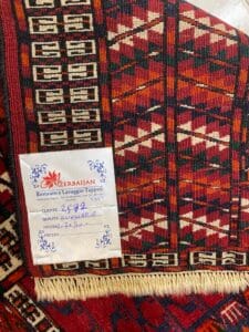 Azerbaijan Tappeti Milano Un tappeto rosso decorato con un'etichetta di riparazione attaccata a un angolo, che mostra dettagli scritti a mano in italiano.